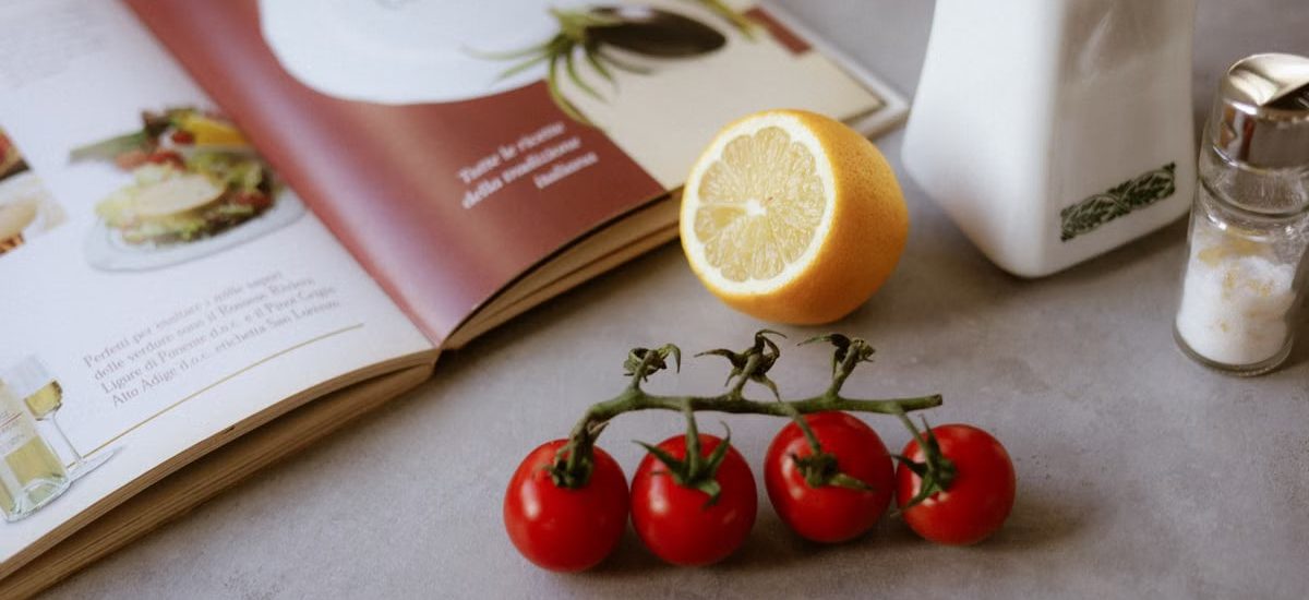 Flat lay of cooking ingredients including tomatoes and a lemon with a cookbook.